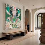 A large framed abstract art print featuring a stylized face with green, teal, pink, and white floral details, displayed on a beige wall in a modern, elegant interior with a bench and arched doorways.