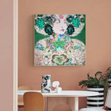 A square framed wall art print featuring a stylized floral portrait of a woman in green and multicolor tones, displayed above a dining table in a peach-colored room with chairs and a plant.