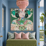 A large framed art print featuring a colorful, ornate portrait with green floral elements and gold detailing, displayed on a blue wall above a green velvet bench with pink pillows in a styled interior.